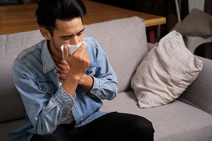 Man holds his chest and blows his nose into a napkin while sitting on a grey couch.